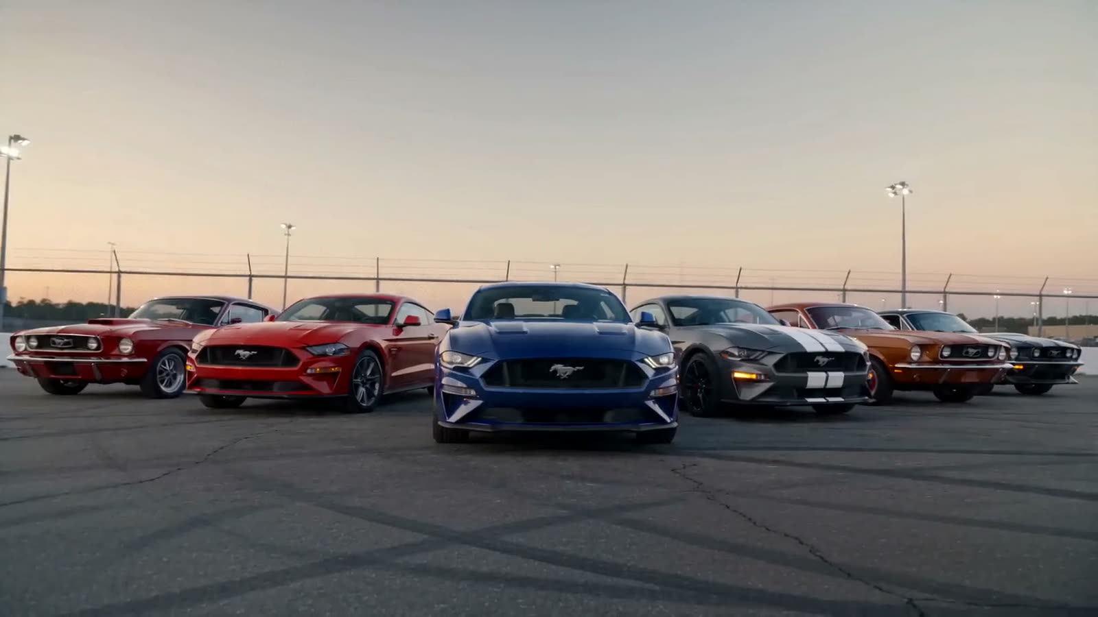 Opening frame of a short film: a row of muscle cars parked nose-forward on a racetrack apron at dusk, floodlights and a chain-link fence behind them.